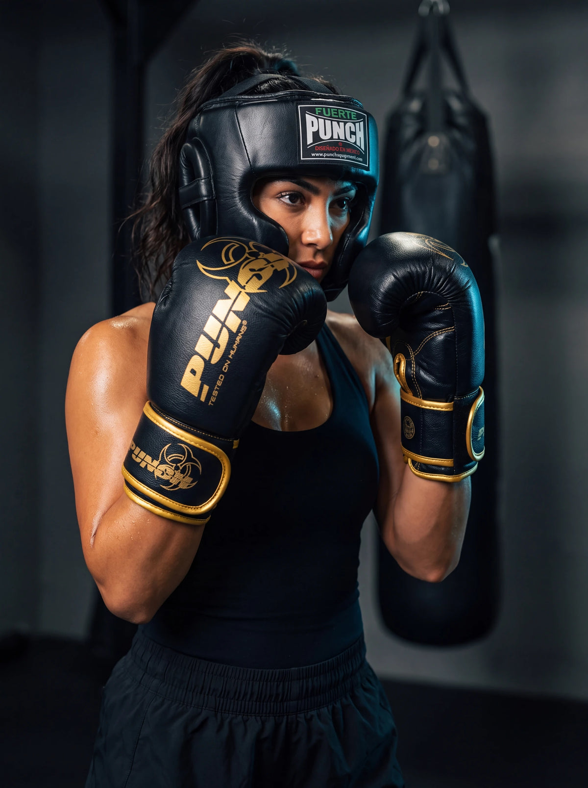 Female boxer wearing the Punch Equipment boxing sparring kit — Fuerte Mexican open-face head gear and black and gold Urban Haymaker 16oz gloves, in guard position in a gym