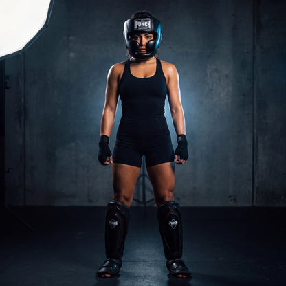 Female fighter wearing the Punch Equipment women's pro protective kit — Trophy Getters full-face head gear and Black Diamond Precision Plus shin pads, standing in a gym