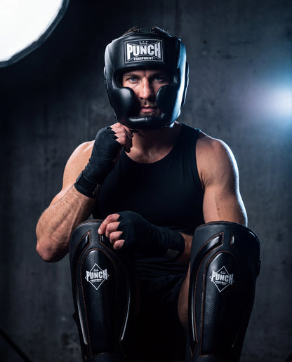 Male fighter wearing the Punch Equipment pro protective kit — Trophy Getters full-face head gear, wrapped hands and Black Diamond Precision Plus shin pads, in a gym
