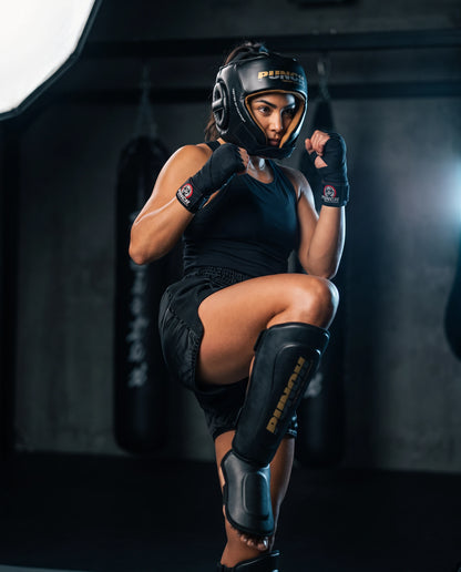Female fighter wearing the Punch Equipment women's Muay Thai sparring kit — Urban open-face head gear, Urban Muay Thai shin pads and wrapped hands, checking a kick in a gym