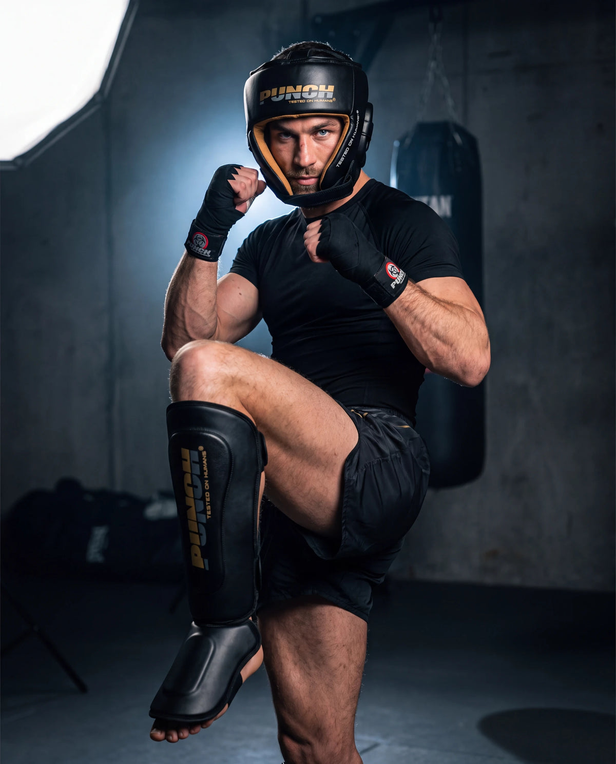 Male fighter wearing the Punch Equipment Muay Thai sparring kit — Urban open-face head gear, Urban Muay Thai shin pads and wrapped hands, checking a kick in a gym
