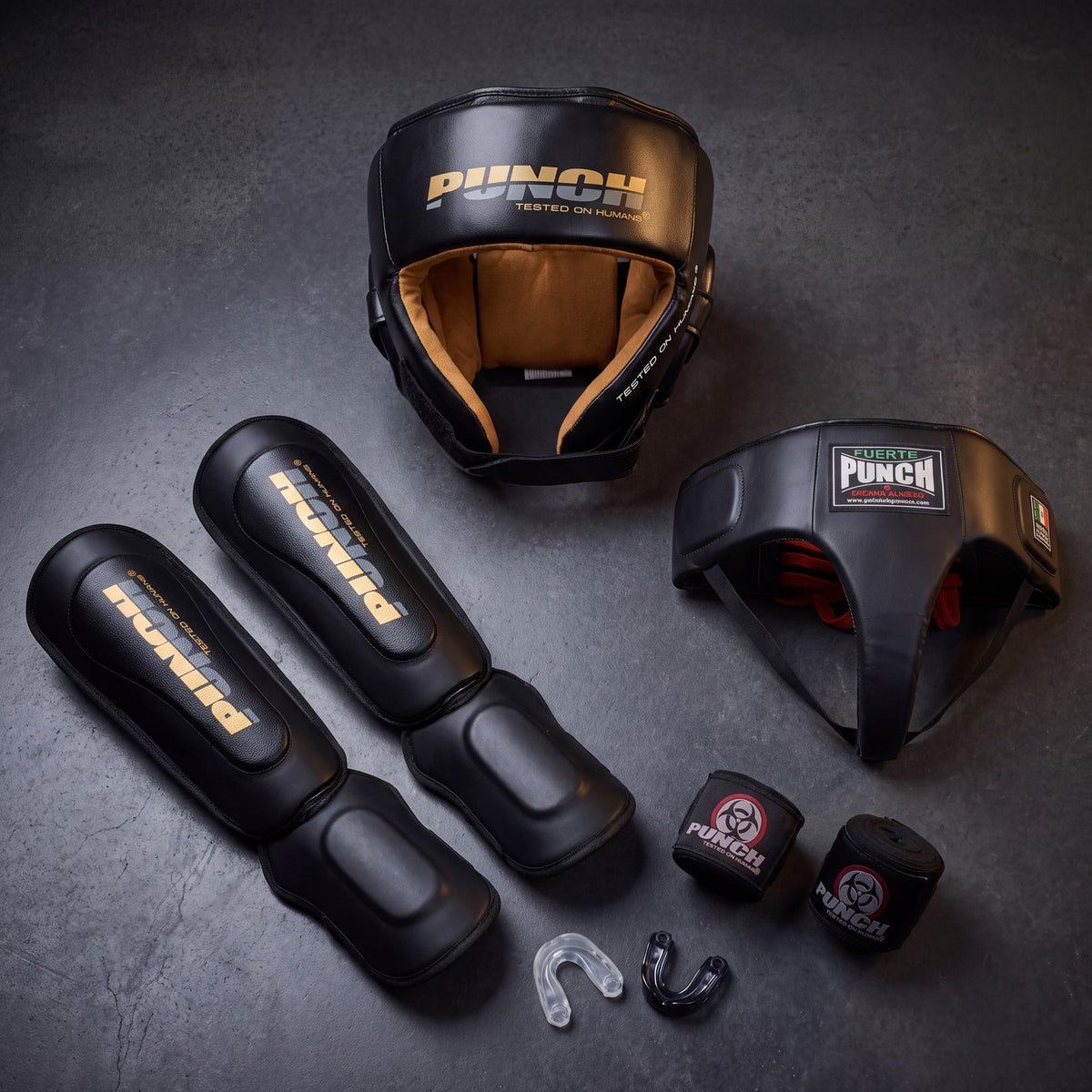 Punch Equipment women's Muay Thai sparring kit flat lay on dark concrete — Urban open-face head gear with tan suede lining, pair of Urban Muay Thai shin pads with gold logos, Mexican Fuerte ladies groin guard, two rolled hand wraps and two gel mouthguards